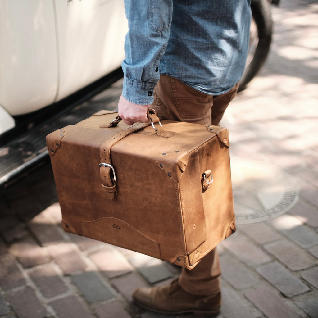 Grandfather Overnight Case in Tobacco carried by its top handle by a man on a brick sidewalk