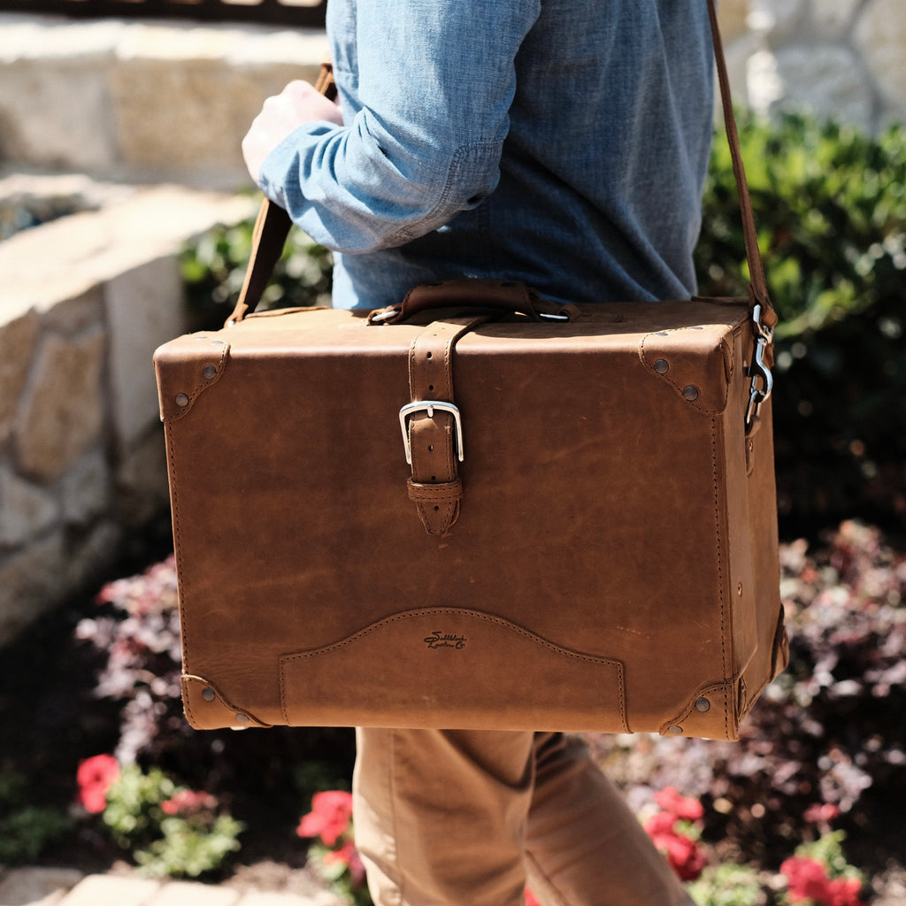 Grandfather Overnight Case in Tobacco worn on the shoulder by a man walking past a stone wall and flowers