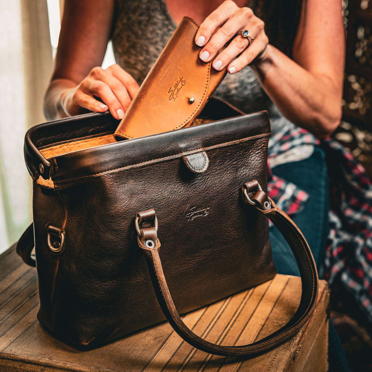 Everyday Purse in Dark Coffee Brown on a wooden stool as a woman tucks a tan leather wallet inside