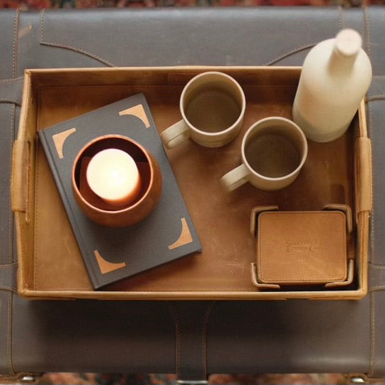 Grandfather Serving Tray in Tobacco leather on an ottoman, holding two mugs, a candle on a book and leather coasters
