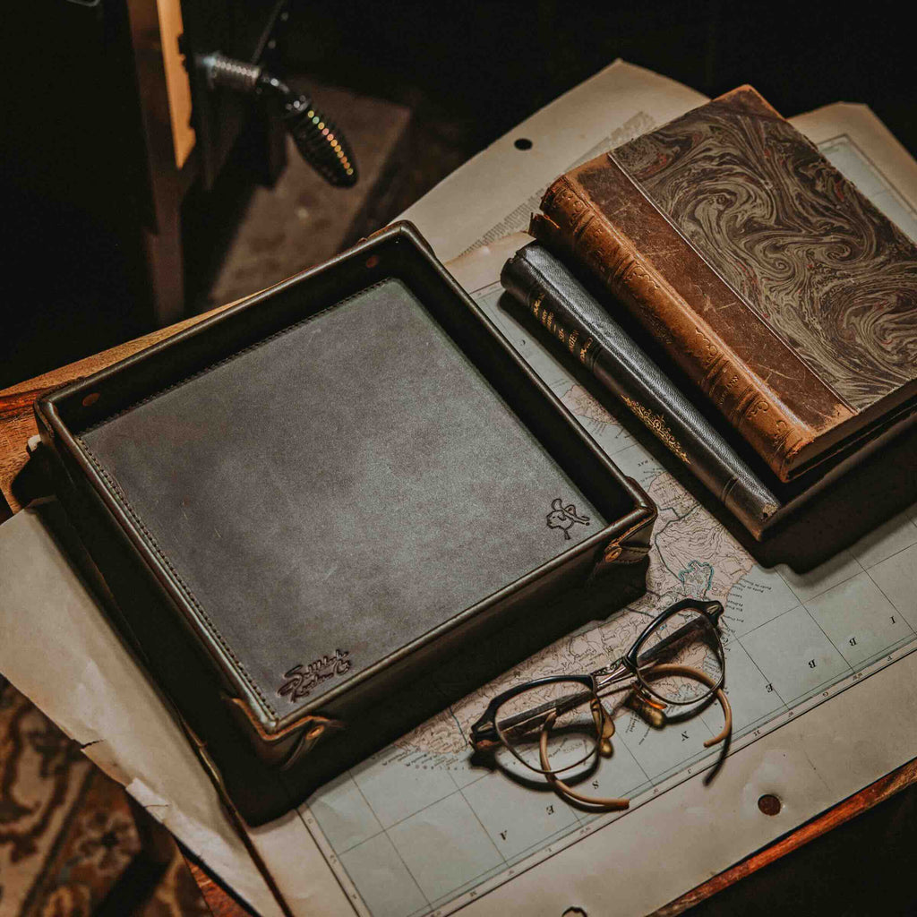 The Leather Grandfather Valet Tray in Dark Coffee Brown on an old map beside marbled books and wire-rim glasses