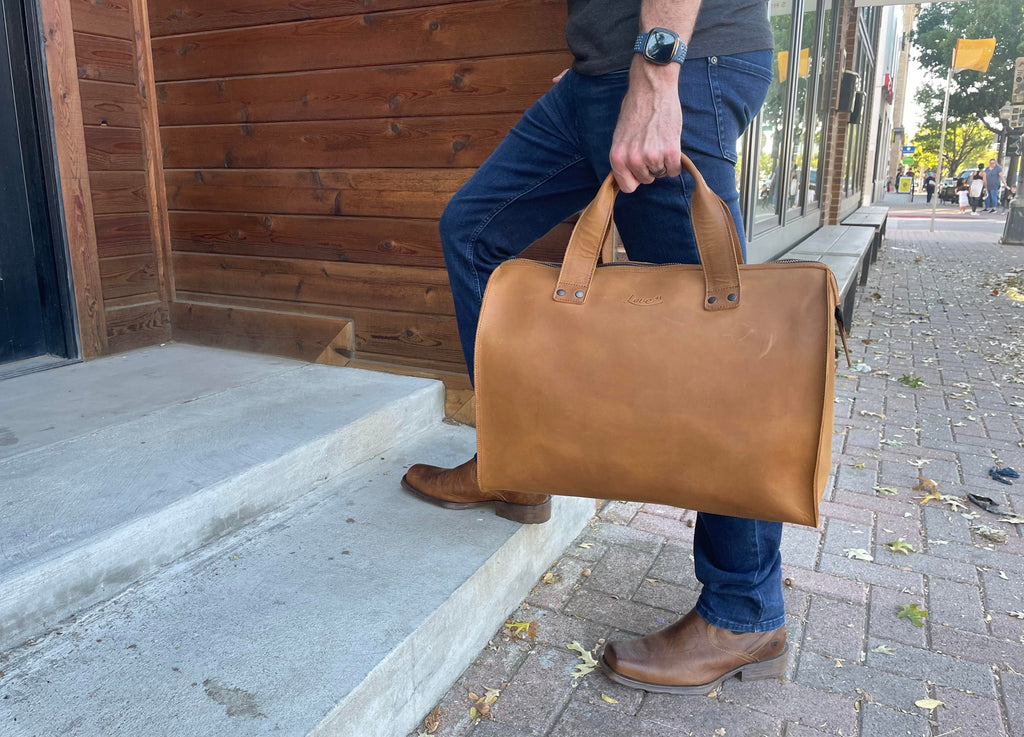 Simple Overnight Leather Duffle Bag in Tobacco, carried by the handles as a man steps onto a sidewalk curb