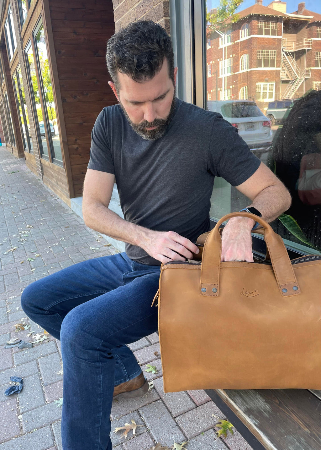 Simple Overnight Leather Duffle Bag in Tobacco, man seated on a sidewalk bench reaching into the open bag
