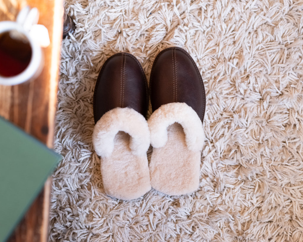 Men's Sheepskin Slippers in Dark Coffee Brown, top-down view of pair on a shaggy rug beside coffee