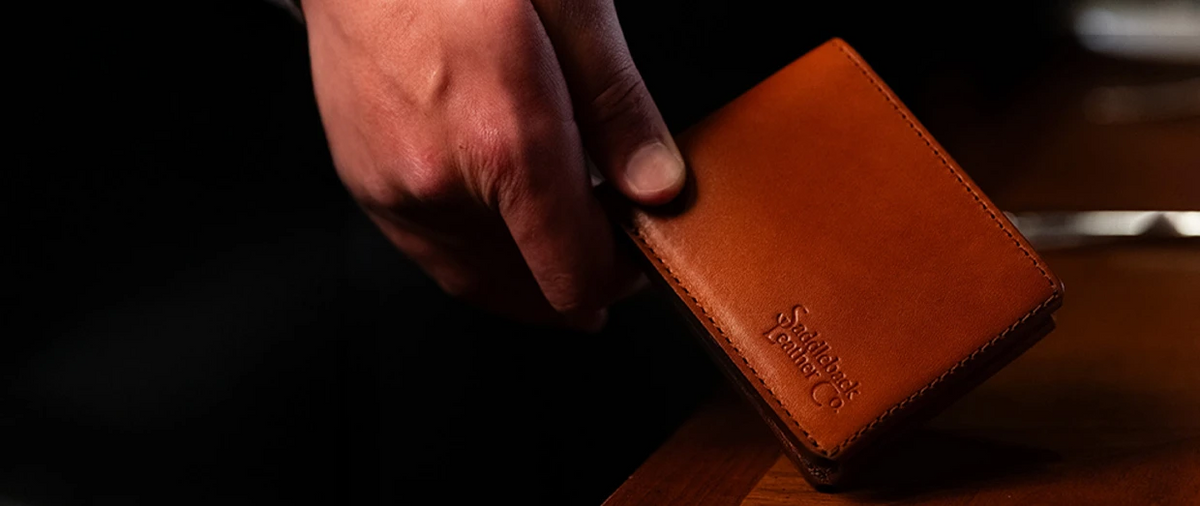 Hand holding a brown leather Penn bifold wallet embossed with the Saddleback Leather Co. logo against a dark background