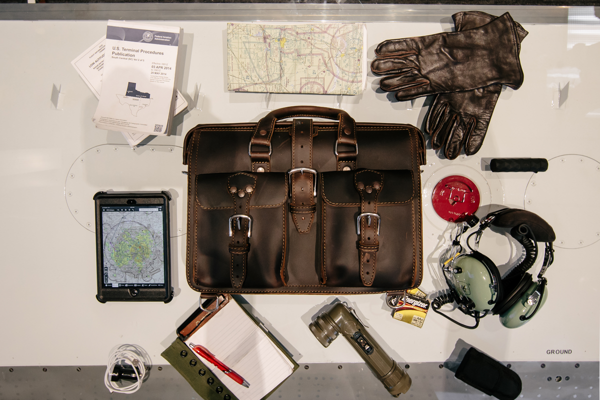 Dark brown Saddleback Leather Flight Bag laid out with pilot gear: gloves, headset, tablet, map, flashlight, and notebook