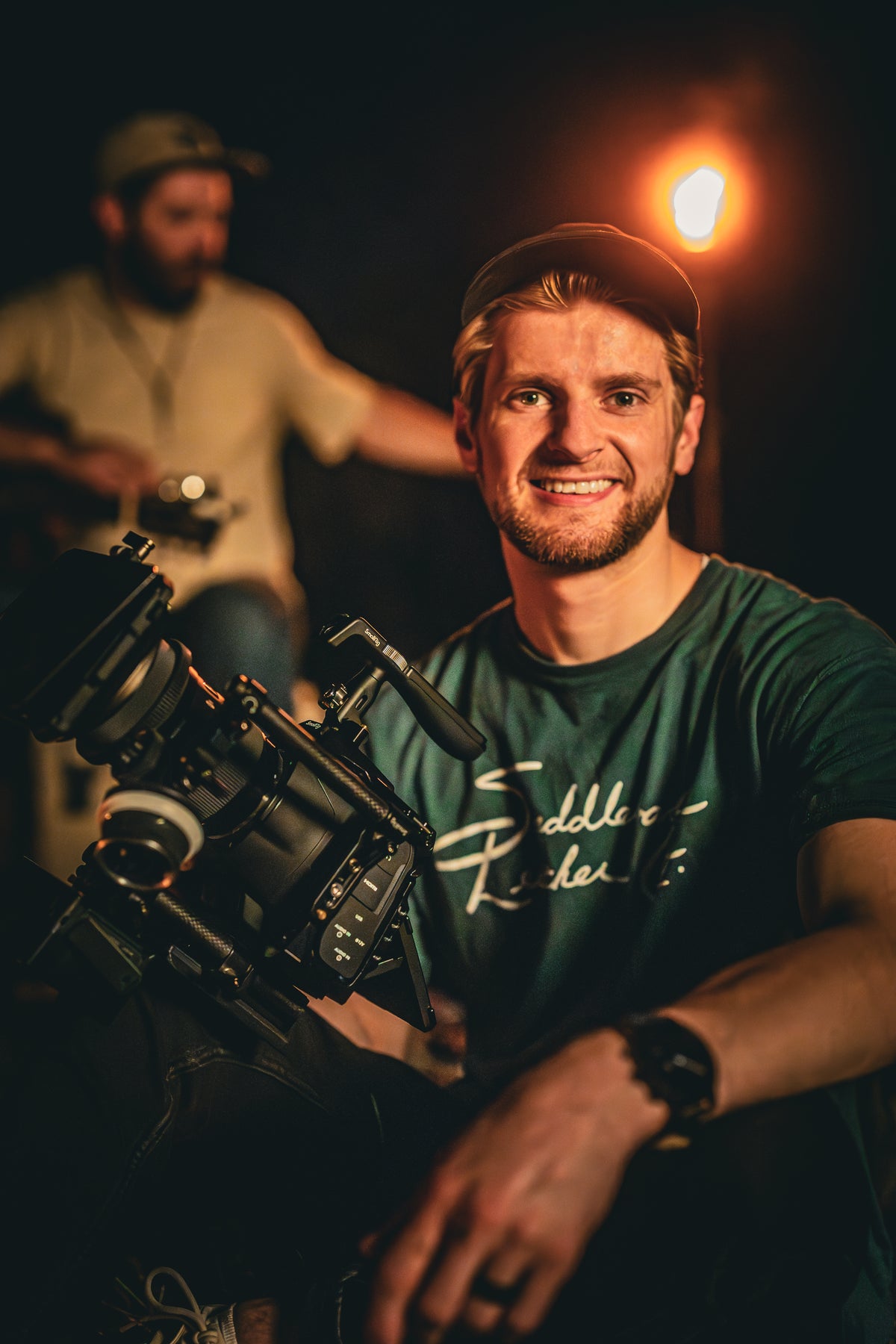 Smiling man in a cap wearing the Signature T-Shirt in Field Green, seated beside a film camera rig