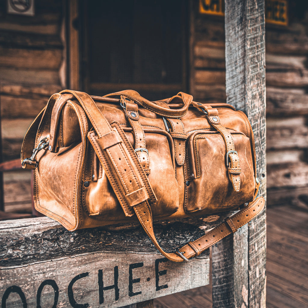 Deep Pocket Leather Duffle Bag in Tobacco resting on a weathered wooden sign outside a rustic cabin