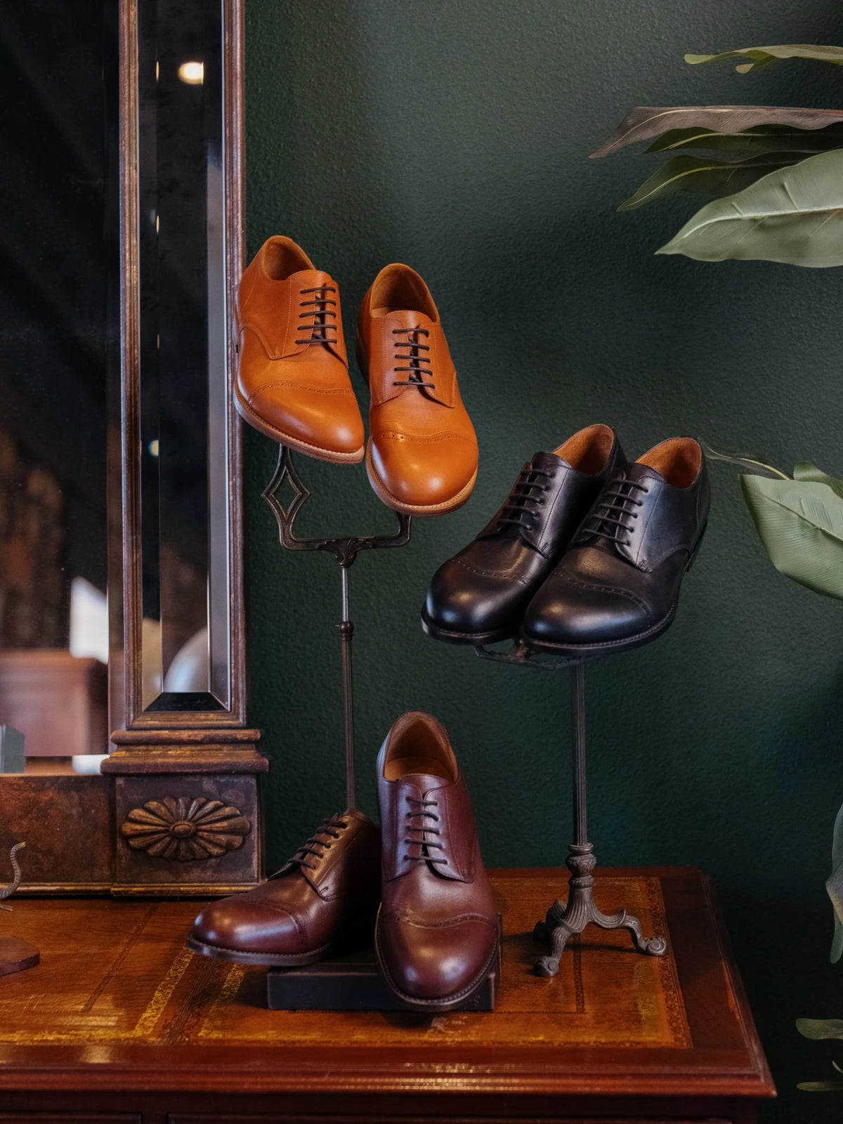 Toe Cap Shoes in English Tan, Raven Black and Dark Oak displayed on stands on an antique desk by a green wall