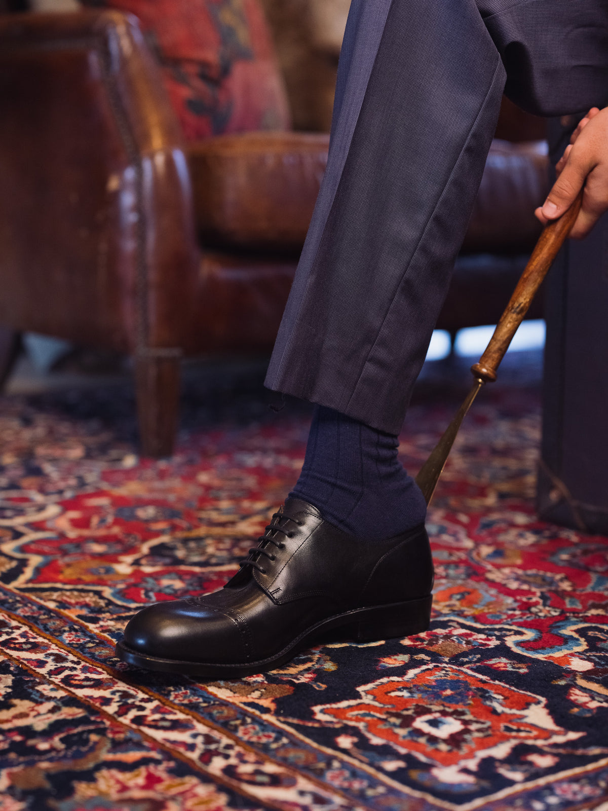 Toe Cap Shoes in Raven Black, single shoe put on with a wooden shoehorn beside a leather armchair