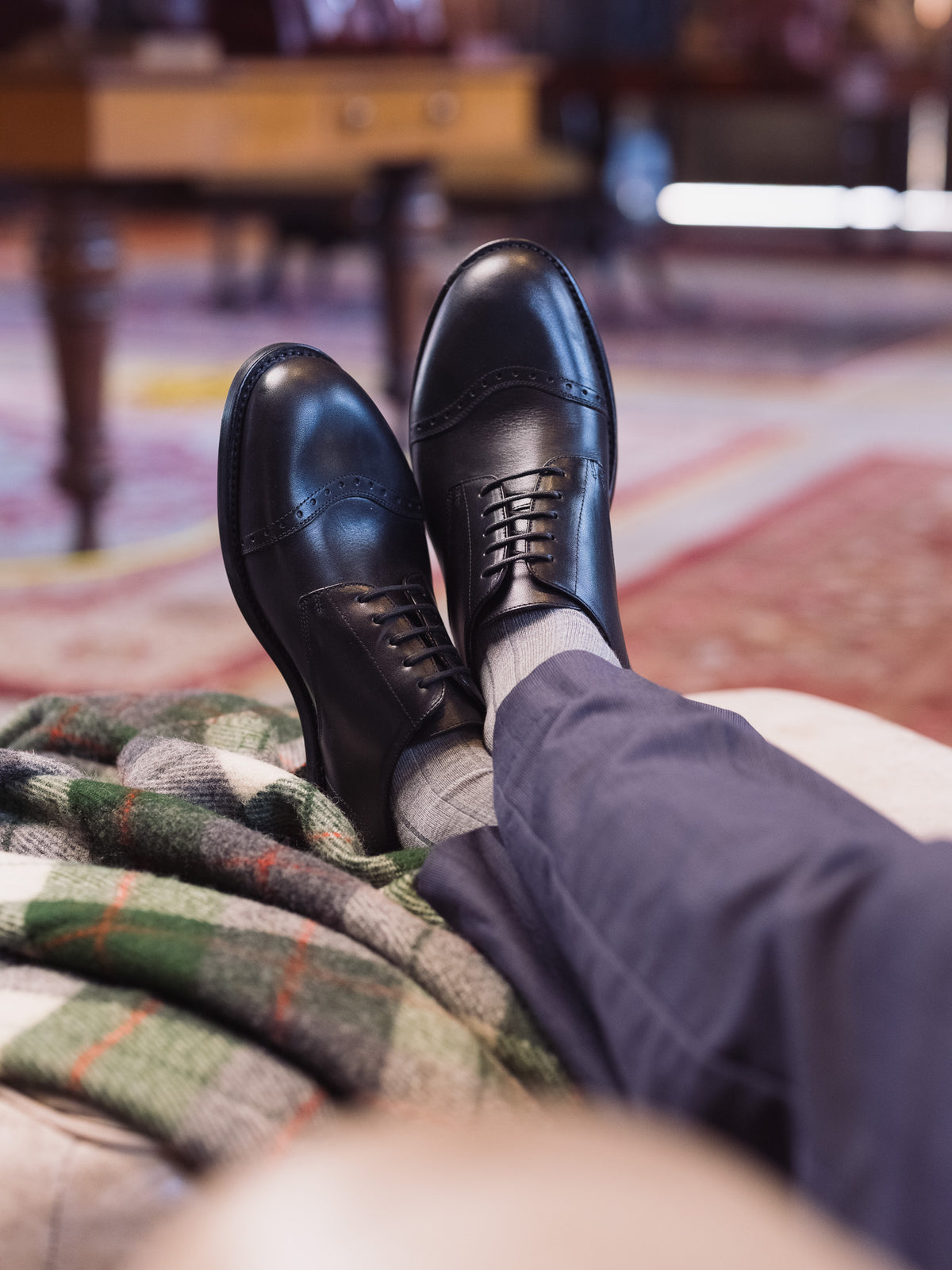 Toe Cap Shoes in Raven Black, worn with gray socks, crossed feet propped on a plaid blanket