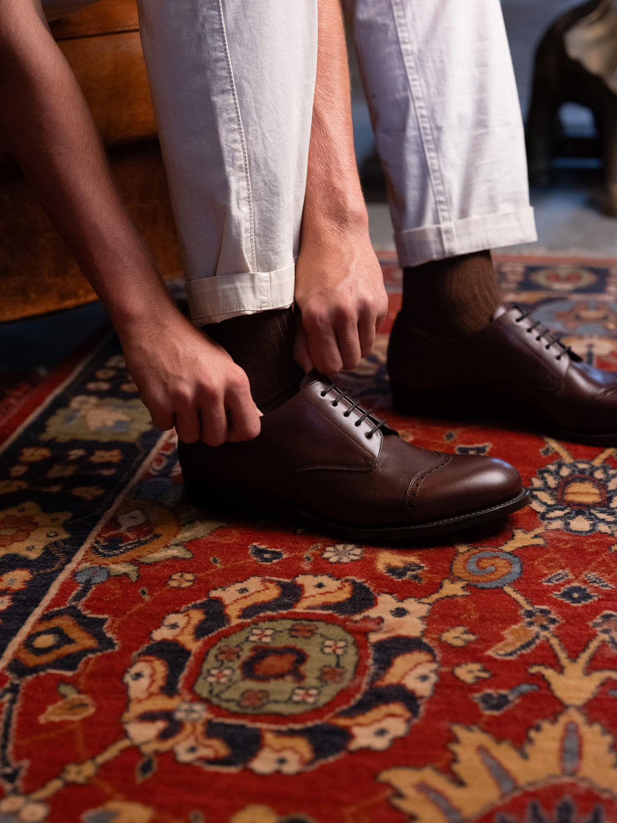 Toe Cap Shoes in Dark Oak, adjusting the laces while seated on a red patterned rug