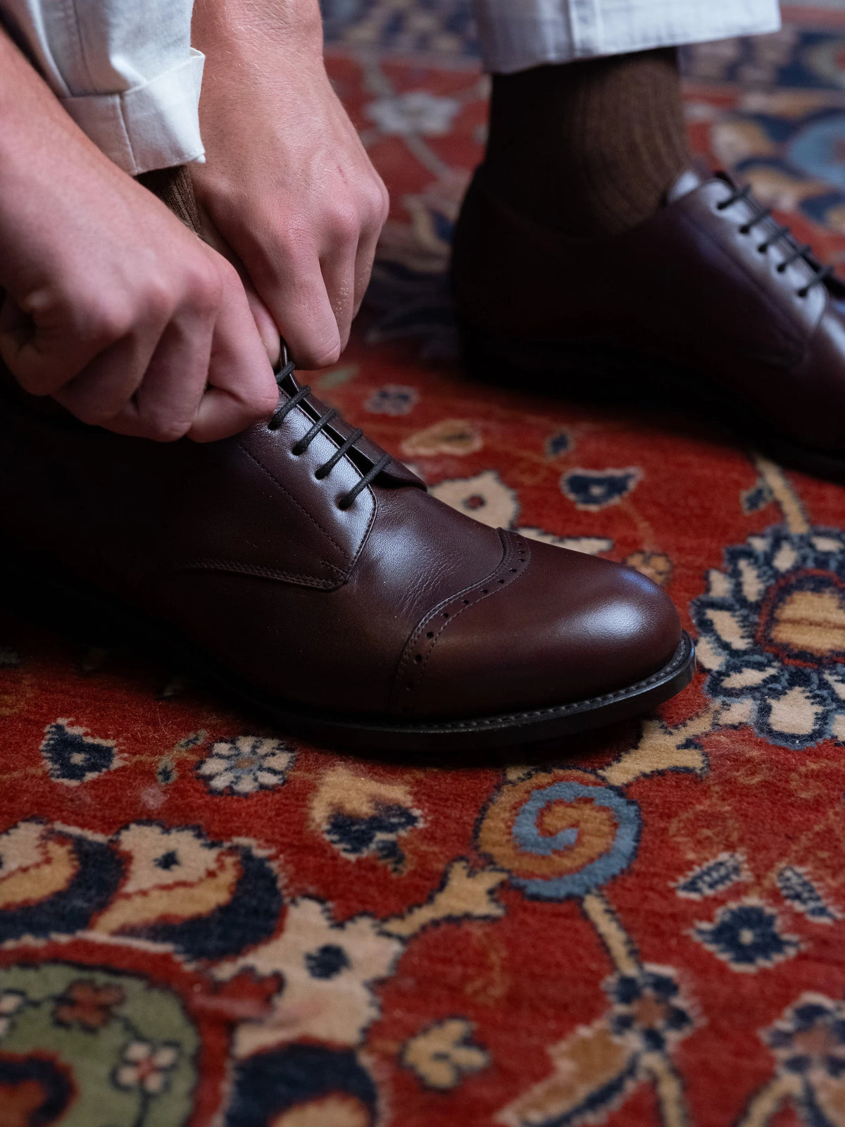 Toe Cap Shoes - Dark Oak, close-up of hands tying the laces on a red patterned rug