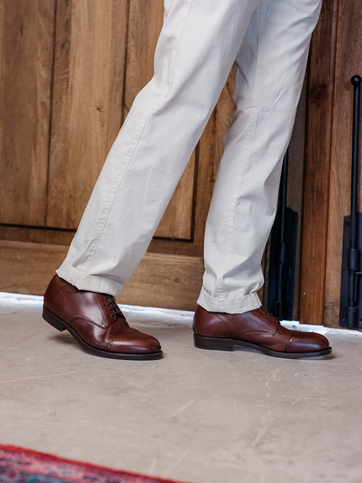 Toe Cap Shoes in Dark Oak, worn with light trousers on a concrete floor by a wooden door