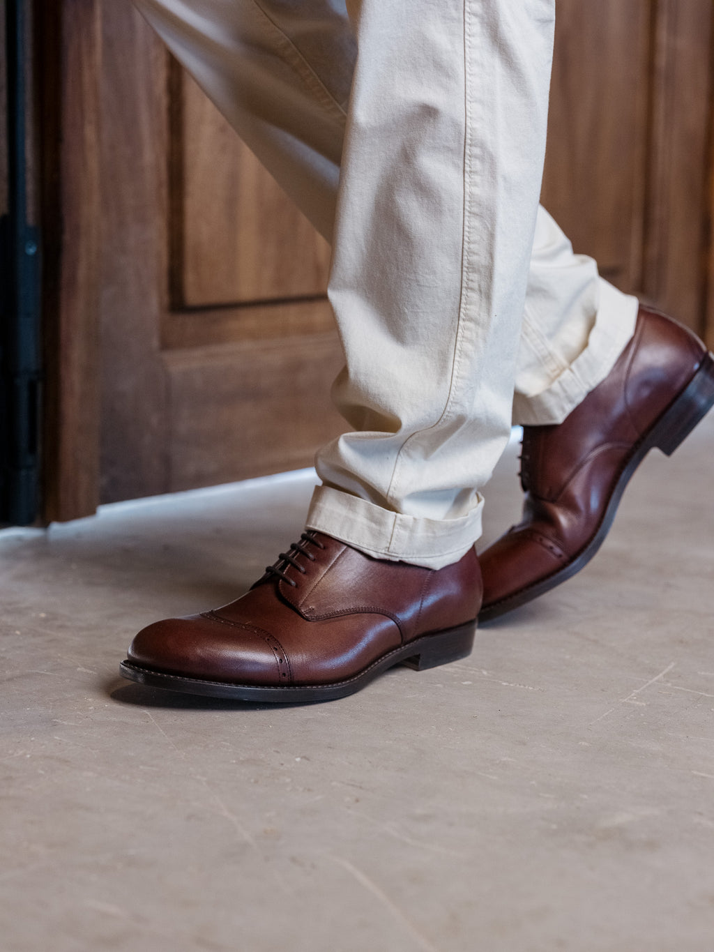 Toe Cap Shoes in Dark Oak, worn walking past a wooden door with cream trousers