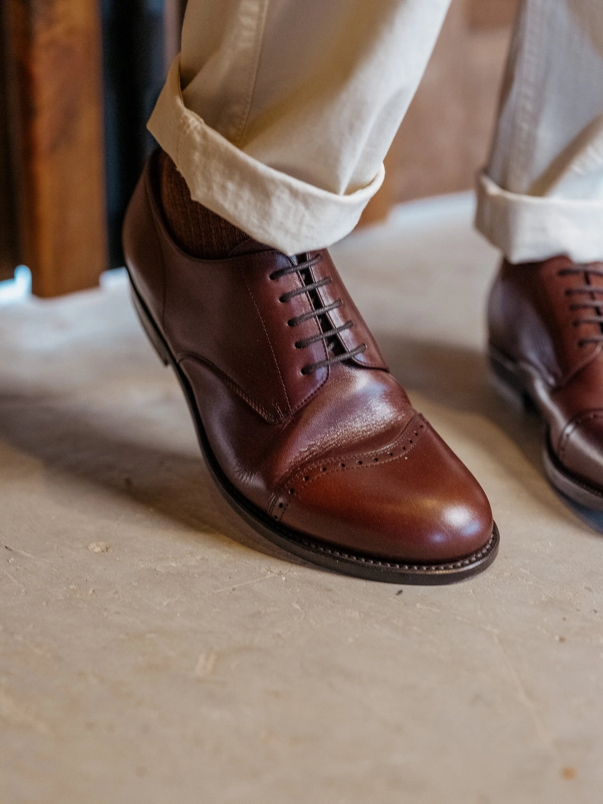 Toe Cap Shoes - Dark Oak on foot, front three-quarter view with cuffed cream trousers on a stone floor