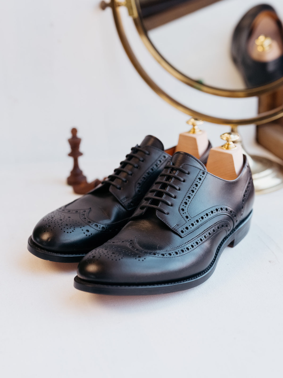 Wingtip Shoes in Raven Black, three-quarter view of the pair with shoe trees and brass mirror