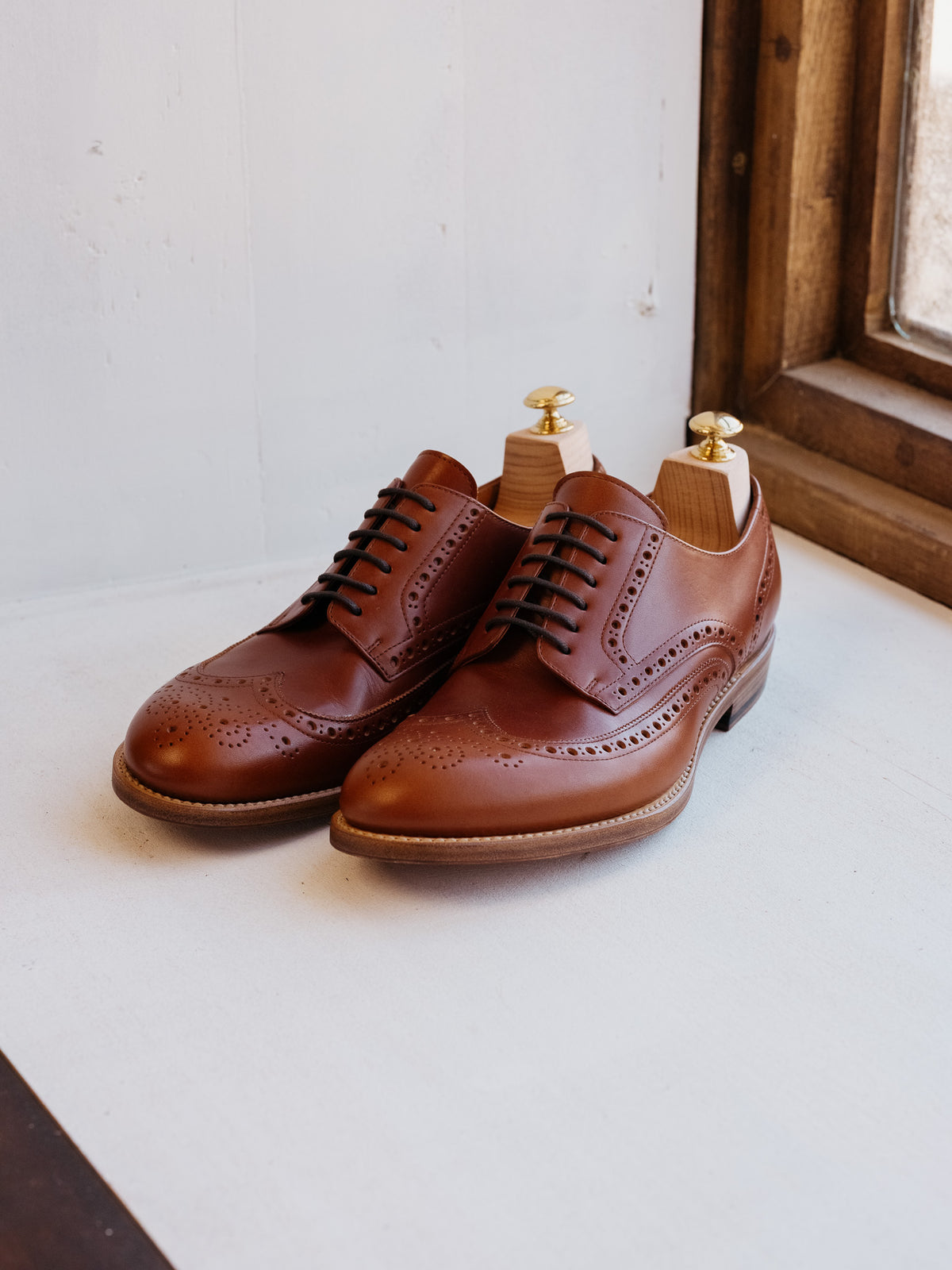 Wingtip Shoes in Chestnut, pair with shoe trees on a white ledge by a wooden window