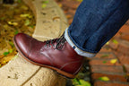 Officer Boot in Oxblood, worn with cuffed jeans, foot resting on a stone ledge with autumn leaves