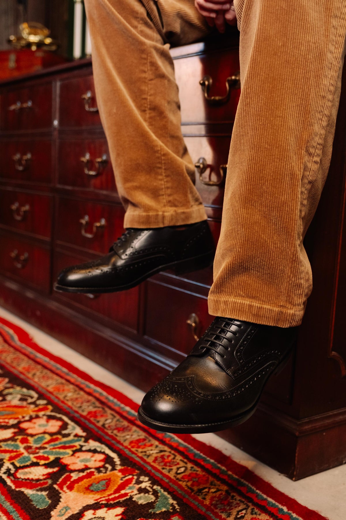 Wingtip Shoes in Raven Black, worn with tan corduroys while seated on a wooden chest