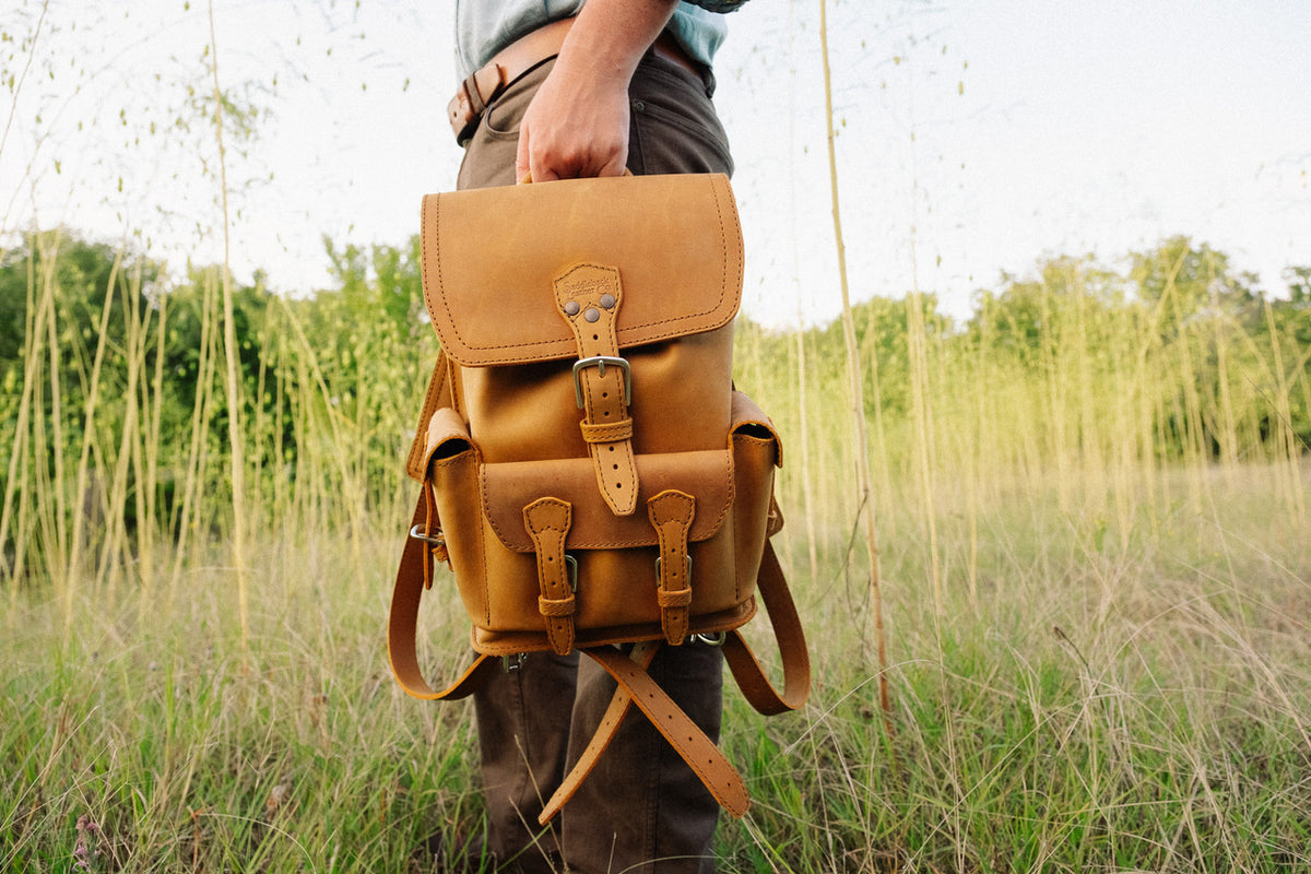 Small Front Pocket Backpack in Tobacco carried by its top handle by a person standing in tall grass