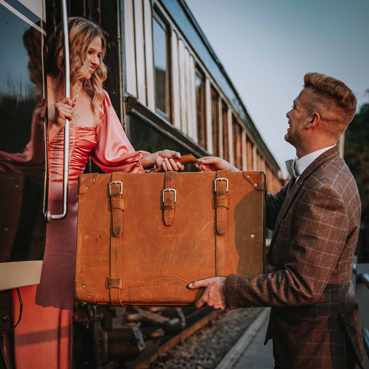 Grandfather Suitcase in Tobacco handed by a man in a tweed suit to a woman boarding a vintage train