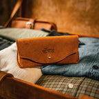 Leather Sunglass Case in Tobacco resting on folded denim and plaid shirts inside an open leather bag