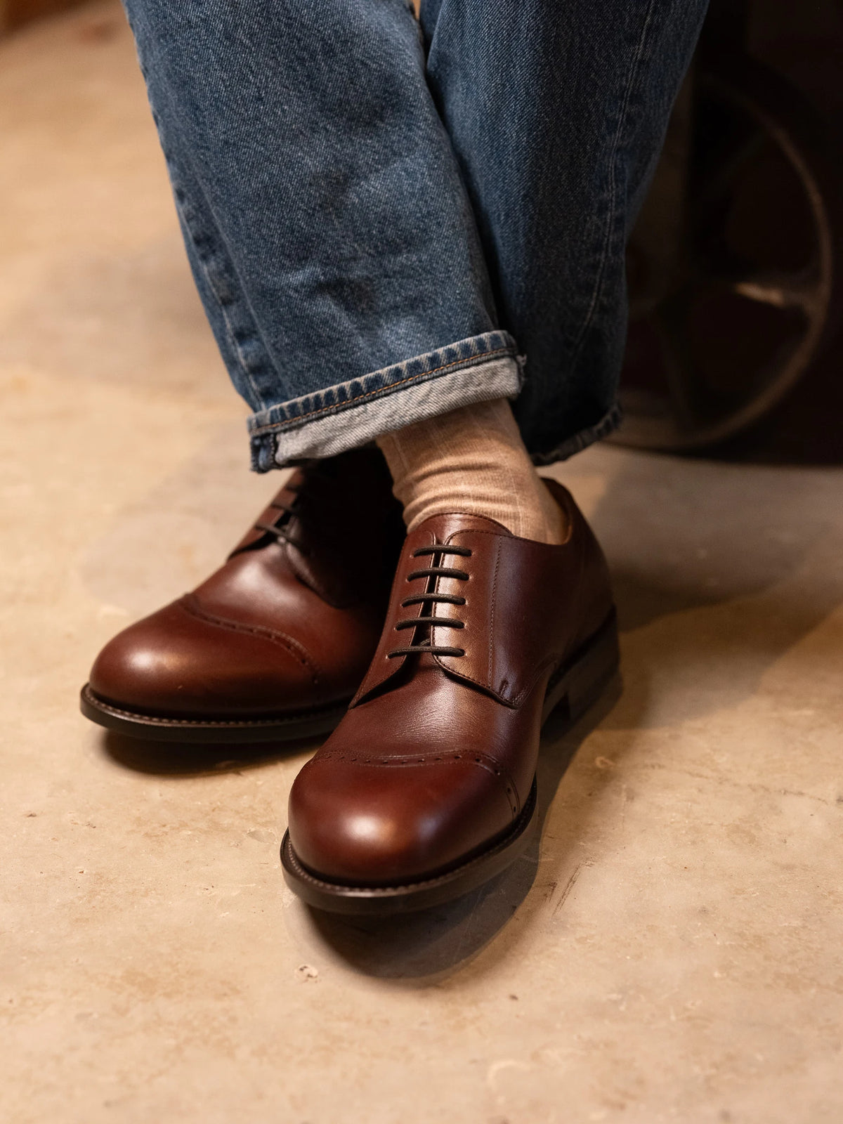 Toe Cap Shoes - Dark Oak worn with cuffed blue jeans and tan socks, crossed ankles on a concrete floor