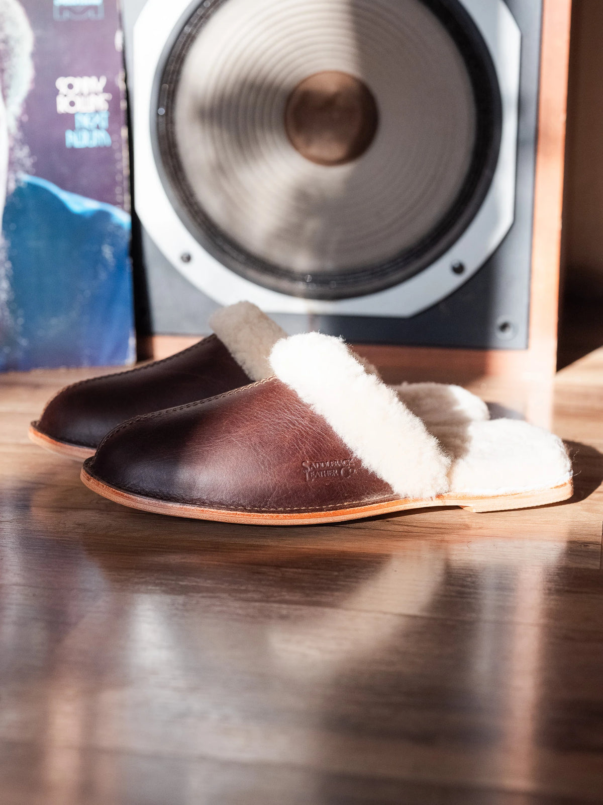 Sheepskin Slippers - Men - New Design in Dark Coffee Brown, side view of a pair on a wood floor in front of a speaker
