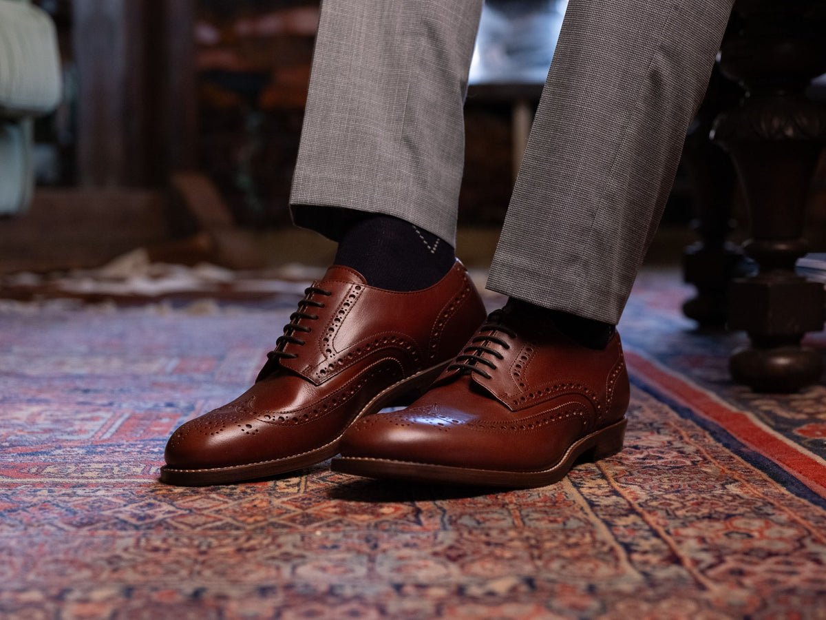 A pair of reddish-brown wingtip brogues worn with grey trousers, standing on a patterned Persian rug.
