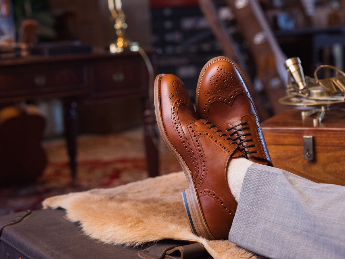 A pair of tan wingtip brogues with the wearer's feet up on a leather trunk, in a study with a writing desk behind.