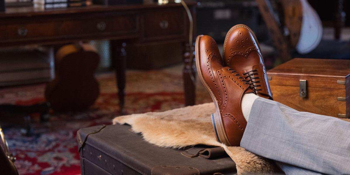 A pair of tan wingtip brogues resting on an old suitcase and a sheepskin, feet up, in a study.