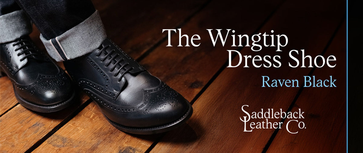 The Wingtip Dress Shoe, Raven Black, Saddleback Leather Co. A pair of black wingtip brogues worn with turned-up grey trousers on a dark wooden floor.