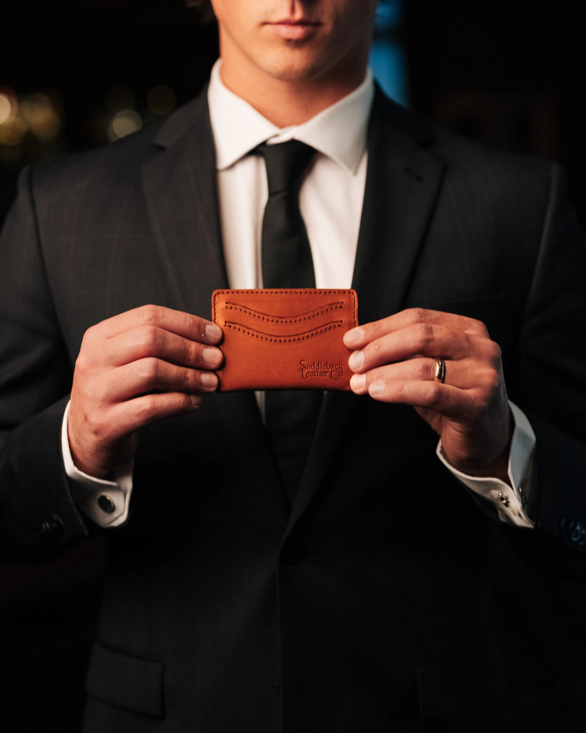 Newton Wallet in English Tan held up in both hands by a man in a dark suit and tie at night