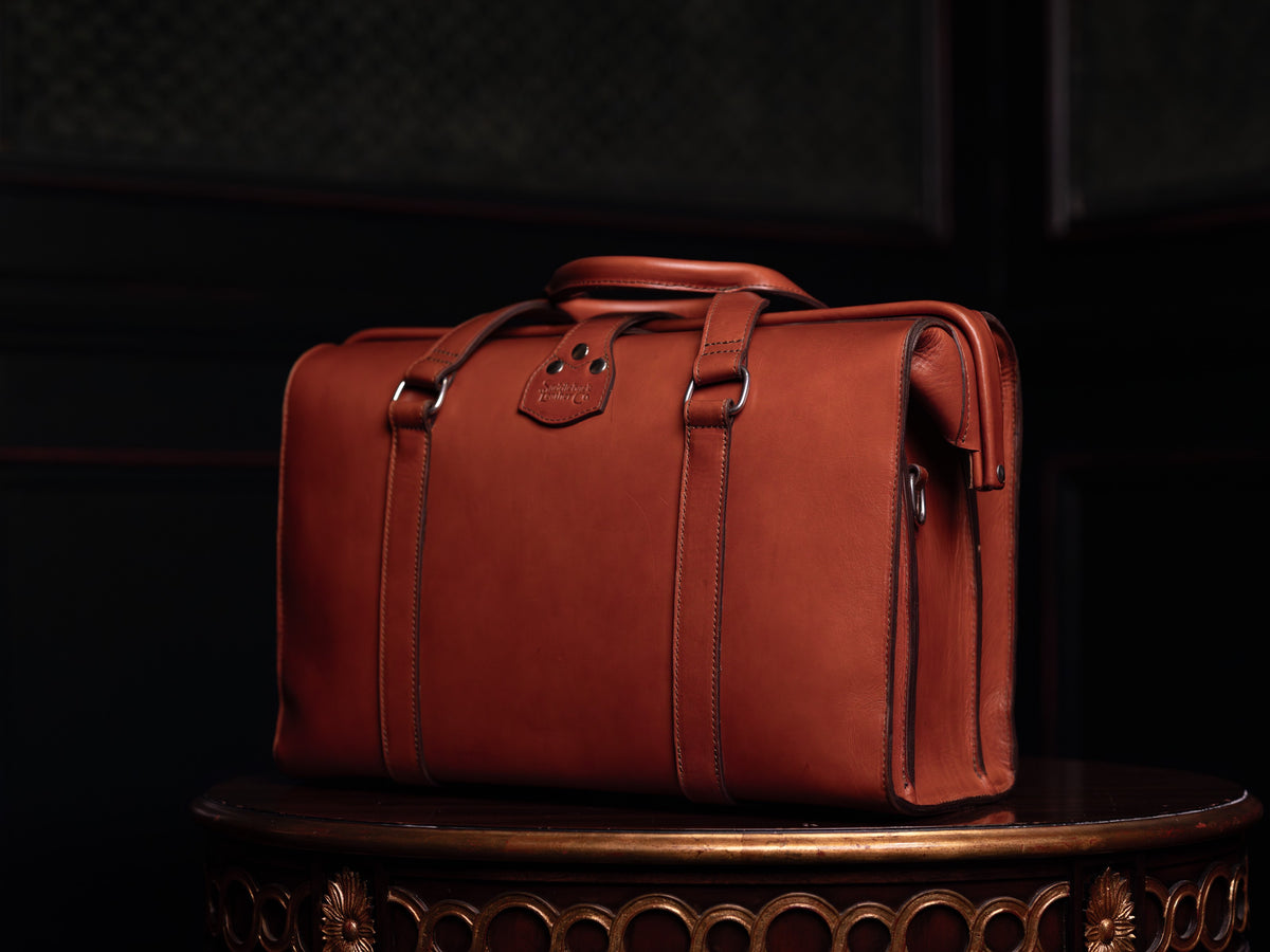Spurgeon Briefcase in English Tan, back view with twin top handles on an ornate wooden pedestal in low light