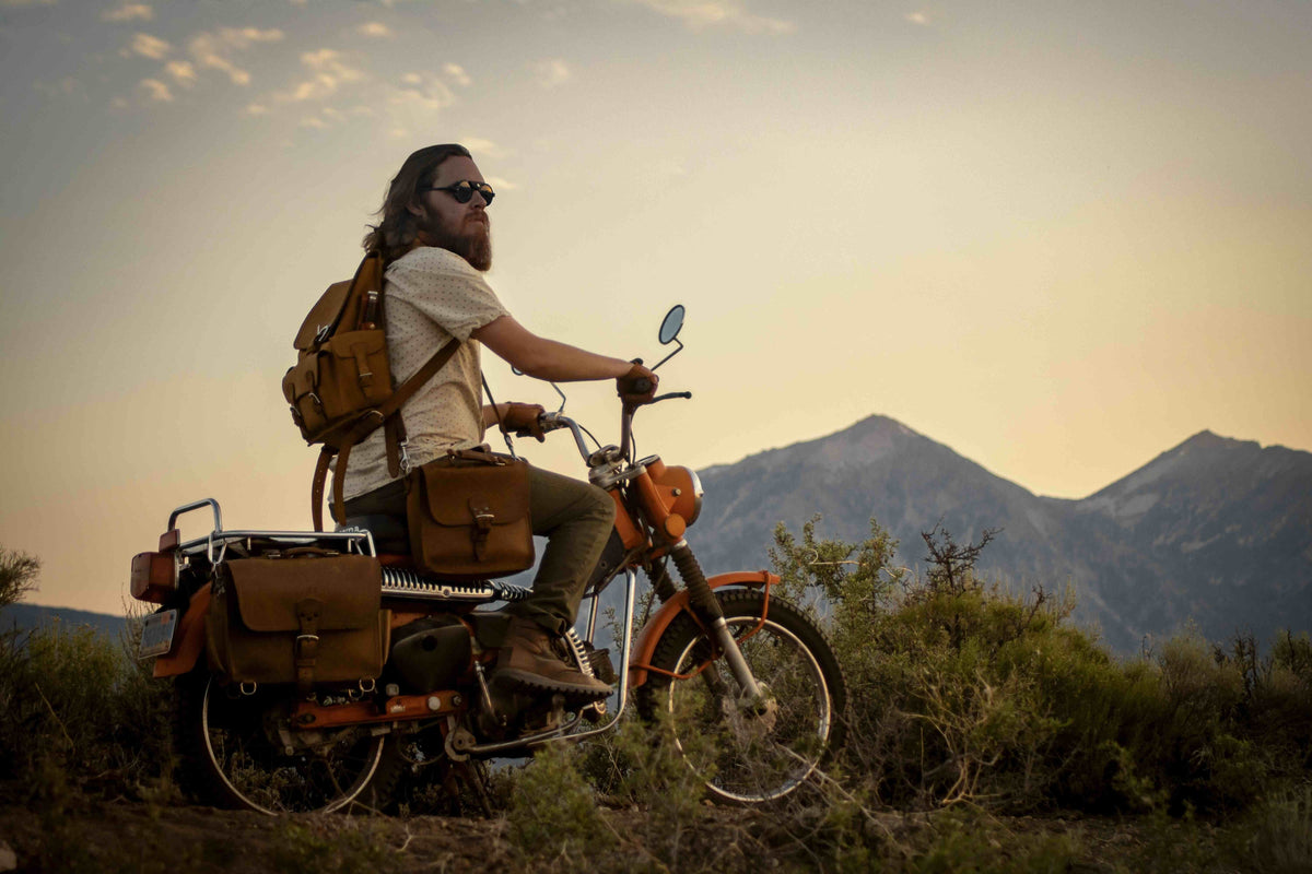 Bearded man on a vintage orange motorcycle with a tan leather slim briefcase and leather bags, mountains at sunset