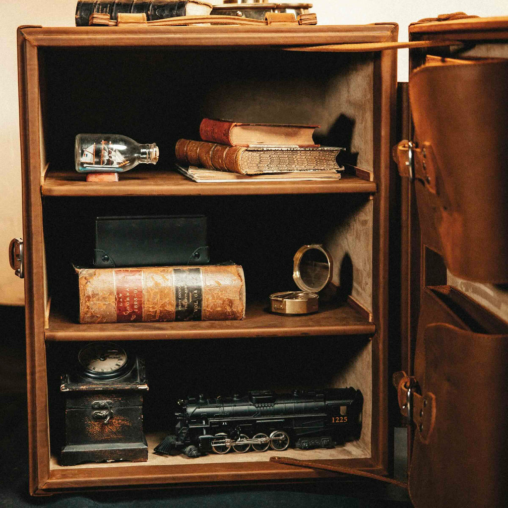 Leather Small Trunk in Tobacco, open upright with shelves holding old books, a ship in a bottle and a model train
