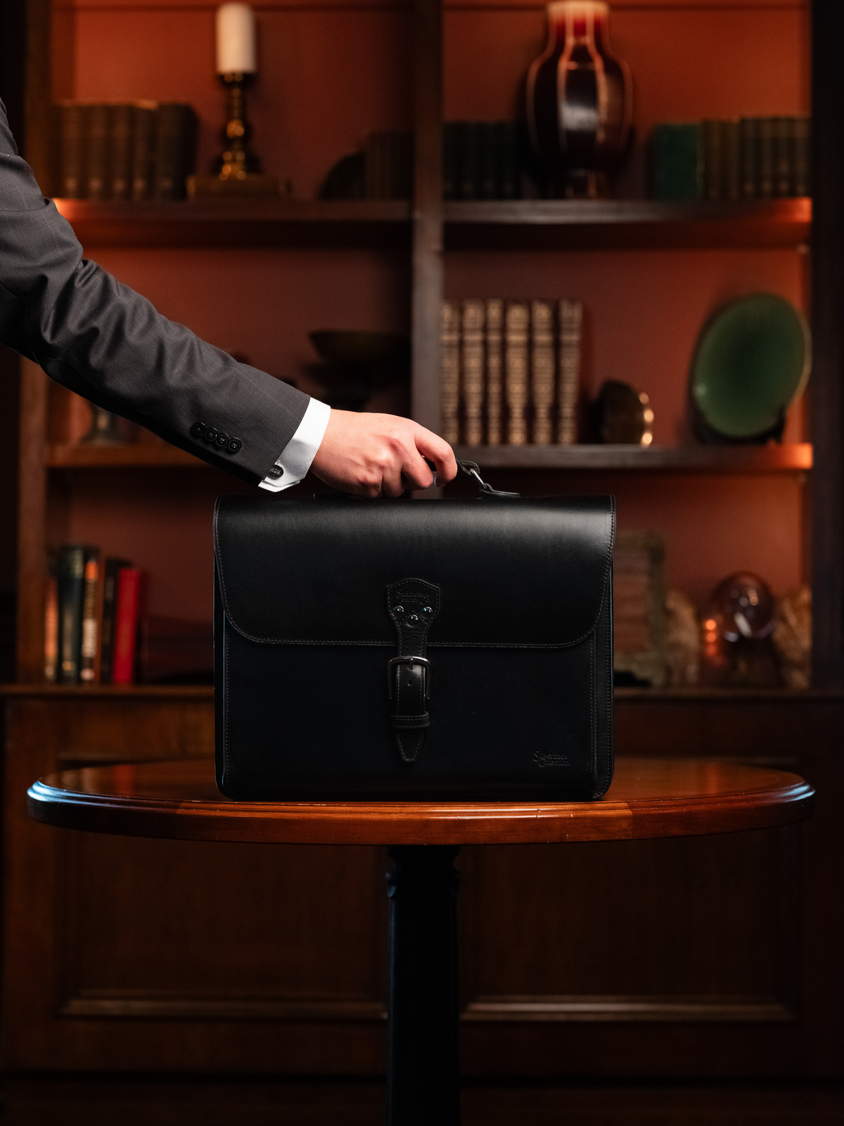 Hand in a suit sleeve holding a black Silverback Livingstone leather briefcase on a round table in a wood-paneled study