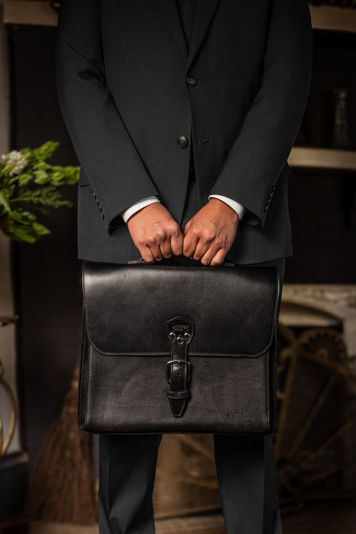 Man in a charcoal suit holding a black leather Livingstone briefcase with a front flap and buckle closure