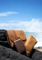 Cash & Receipt Bag in Tobacco on black lava rocks beside other leather goods, ocean waves splashing behind