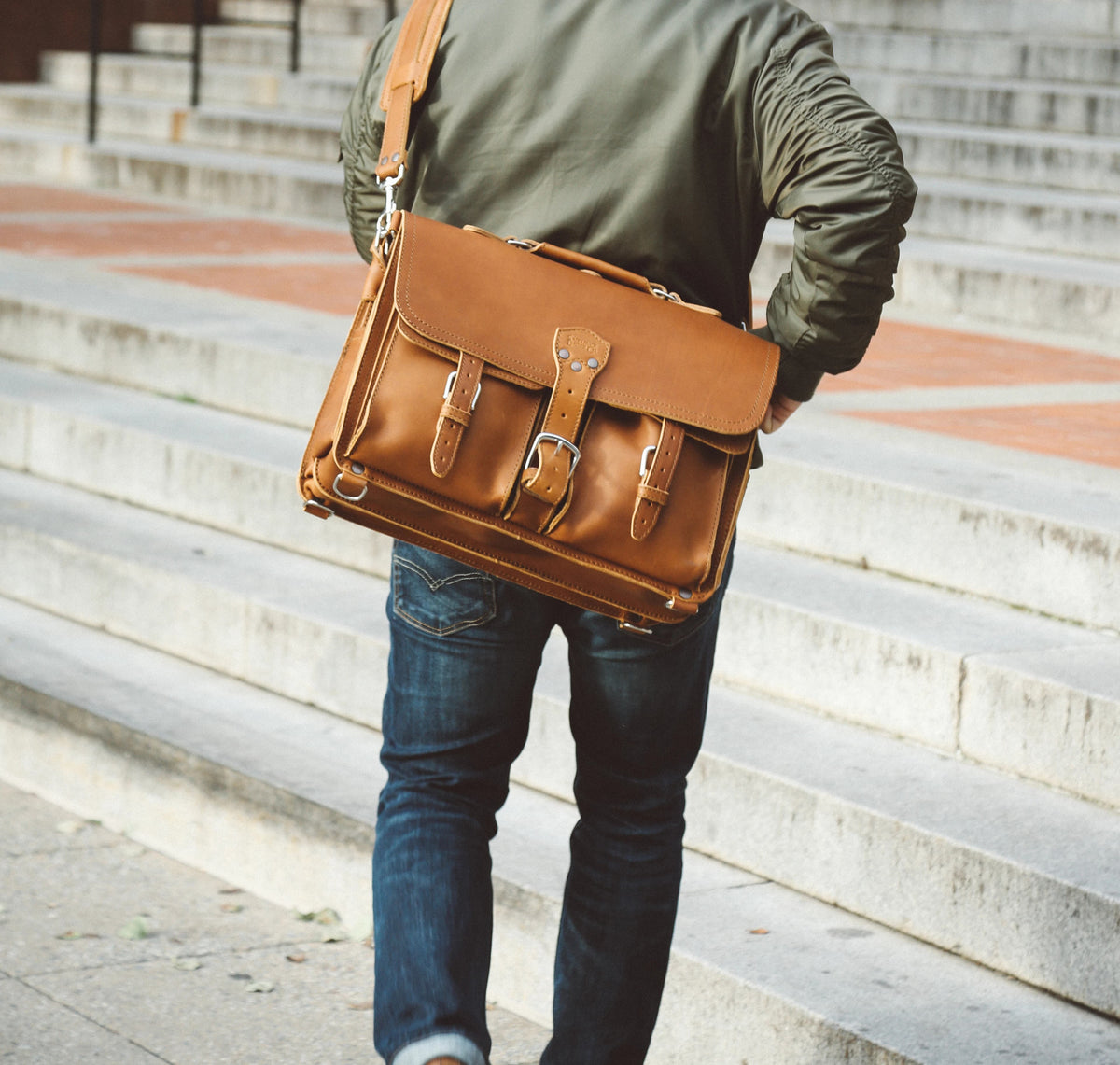 Man in a green jacket walking up concrete steps carrying a tan full grain leather front pocket briefcase