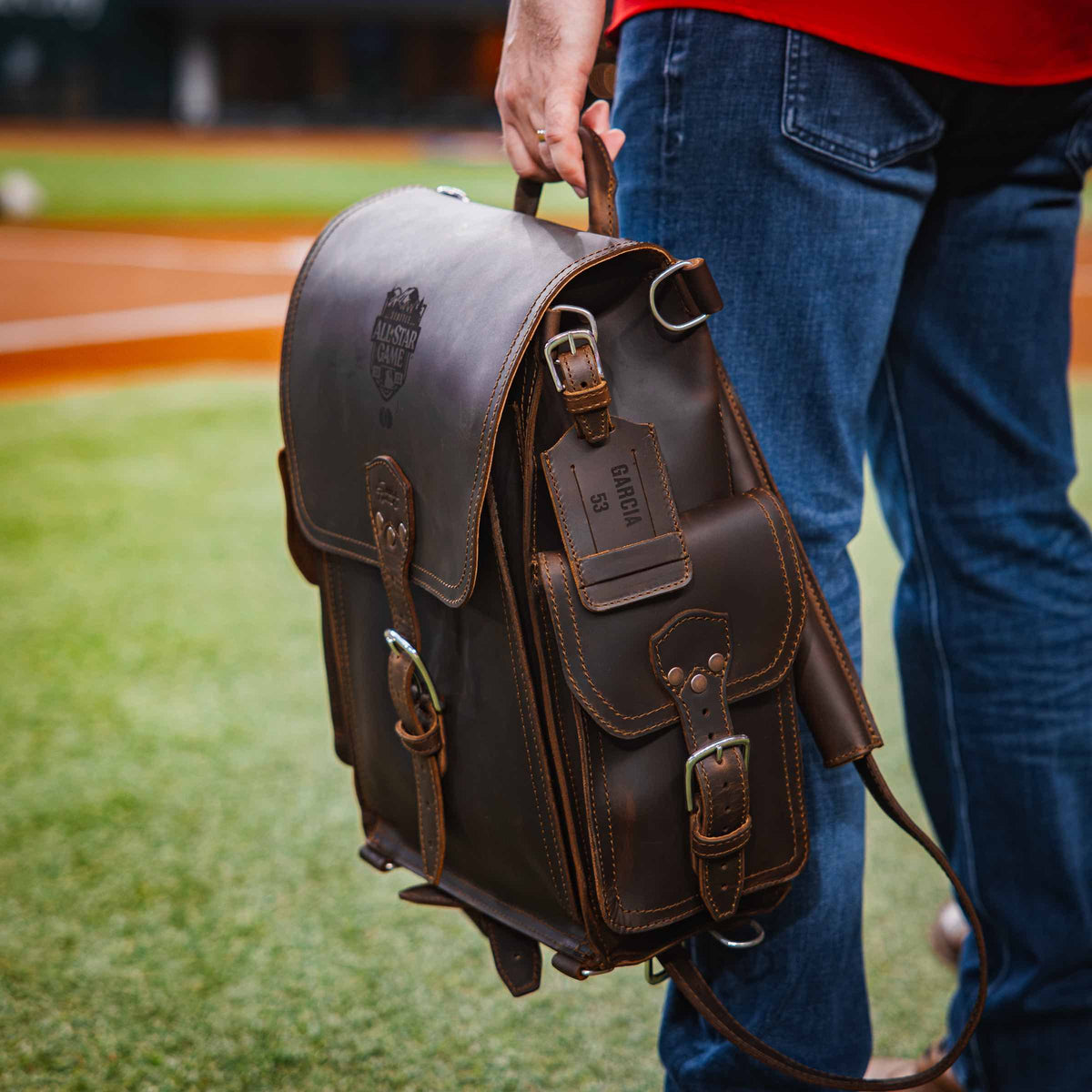 A man on the field holding a dark brown Tank Leather Backpack by its handle, a leather luggage tag clipped to it.