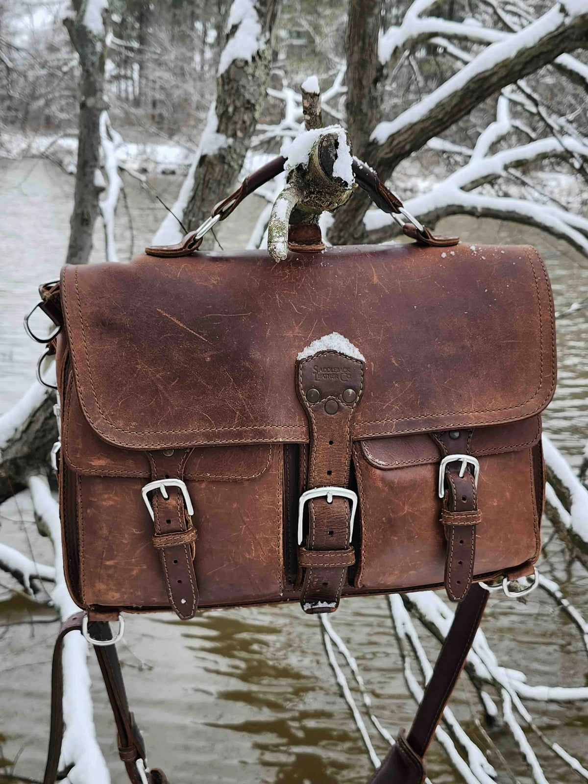 Thin Front Pocket Leather Briefcase in Chestnut with a worn patina, hanging from a snowy branch by a creek