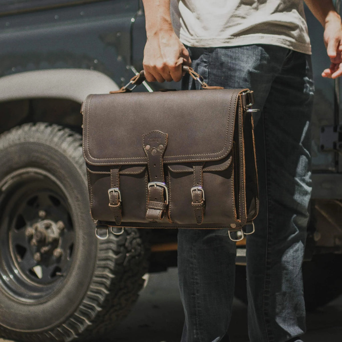 Thin Front Pocket Leather Briefcase in Dark Coffee Brown carried by the handle beside an off-road truck tire