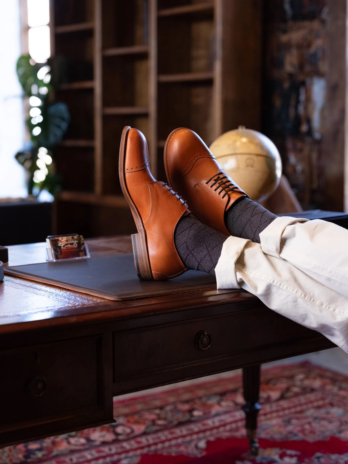 Toe Cap Shoes - English Tan worn by a person with feet up on a wooden desk in a library with a globe