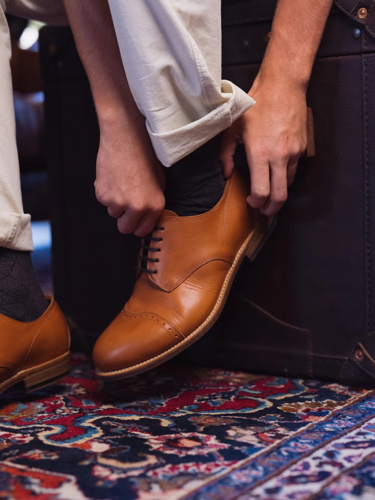 Toe Cap Shoes - English Tan on foot, a person lacing one up while standing on a colorful patterned rug