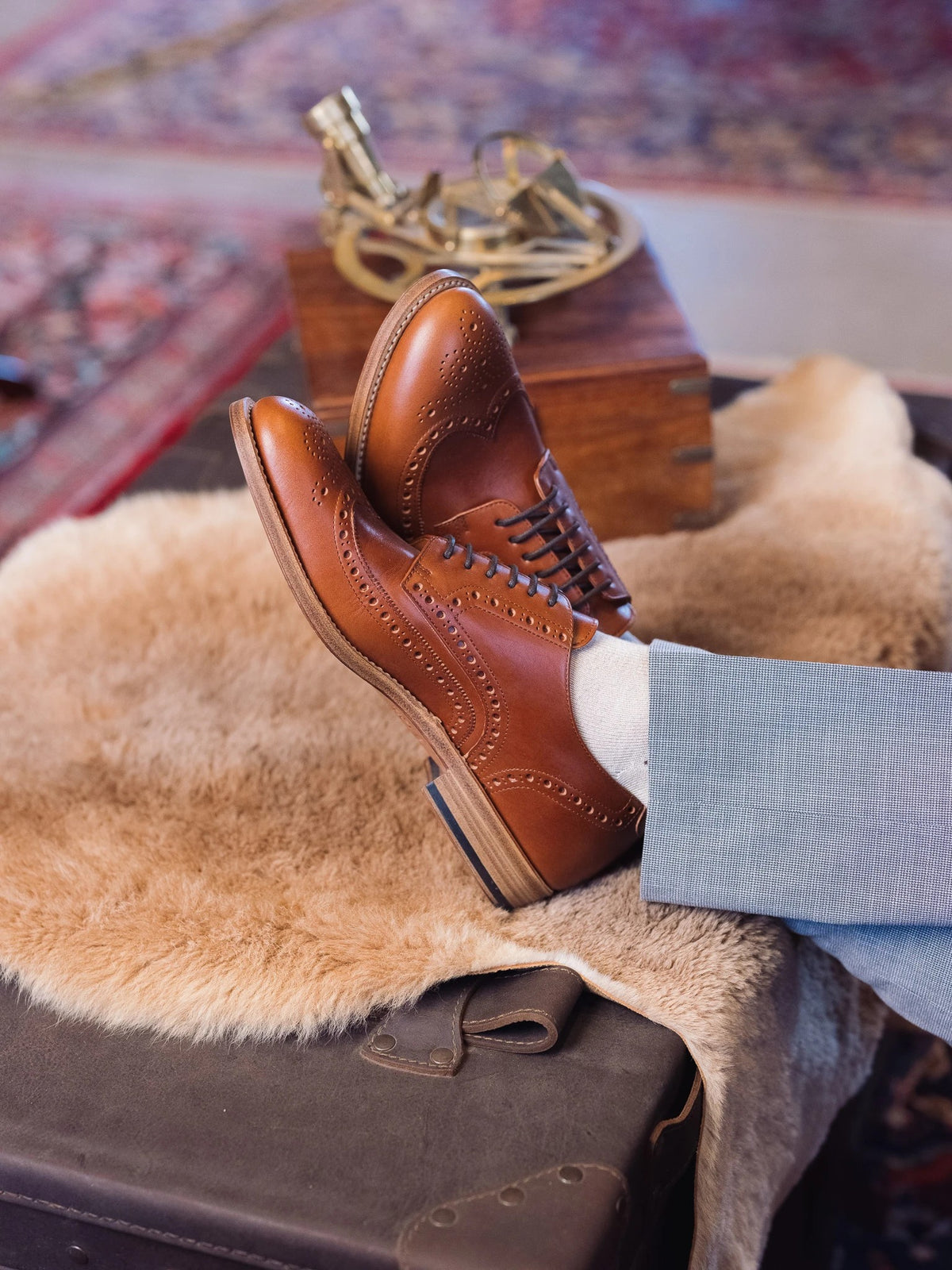 Wingtip Shoes - Natural Tan worn with crossed feet up on a sheepskin-draped trunk beside a brass sextant