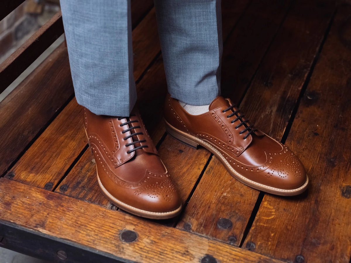 Wingtip Shoes - Natural Tan on foot, a person standing on rustic wood planks in grey trousers