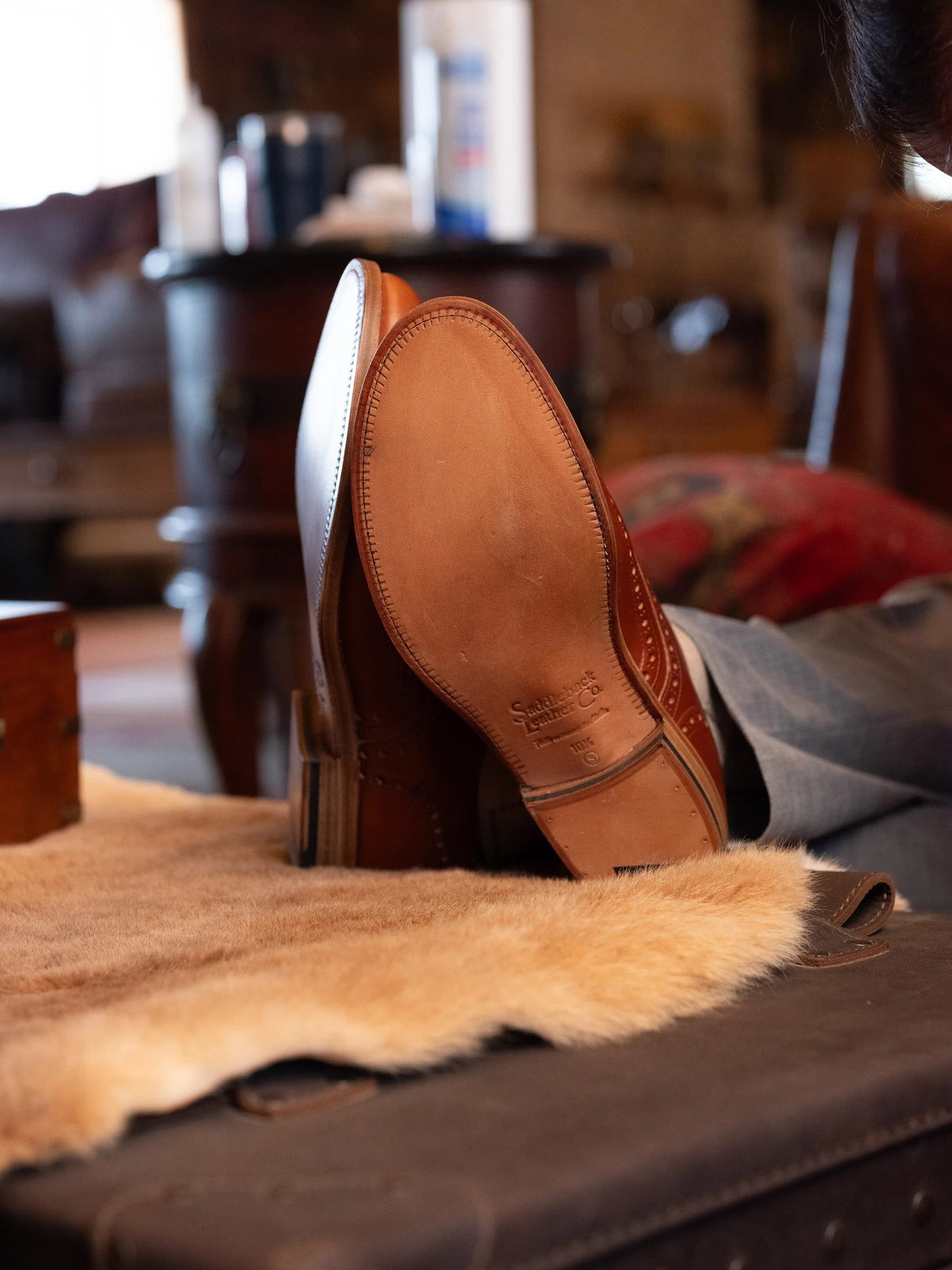 Wingtip Shoes - Natural Tan, leather outsole with stamped logo shown as feet rest on a sheepskin-covered trunk